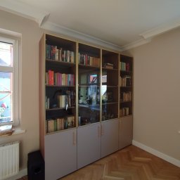 Bauel Tomasz Iskrzak - Zabudowa biblioteczna z przeszklonymi witrynami i szarymi szafkami, widok na parkiet w jodełkę, dekoracyjne listwy przysufitowe. Jasne wnętrze z oknami.