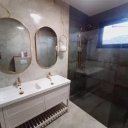 Elegancka łazienka z dwoma umywalkami, złotymi detalami i kabiną prysznicową z szarymi płytkami imitującymi beton. Marmurowe płytki na ścianach i podłodze.