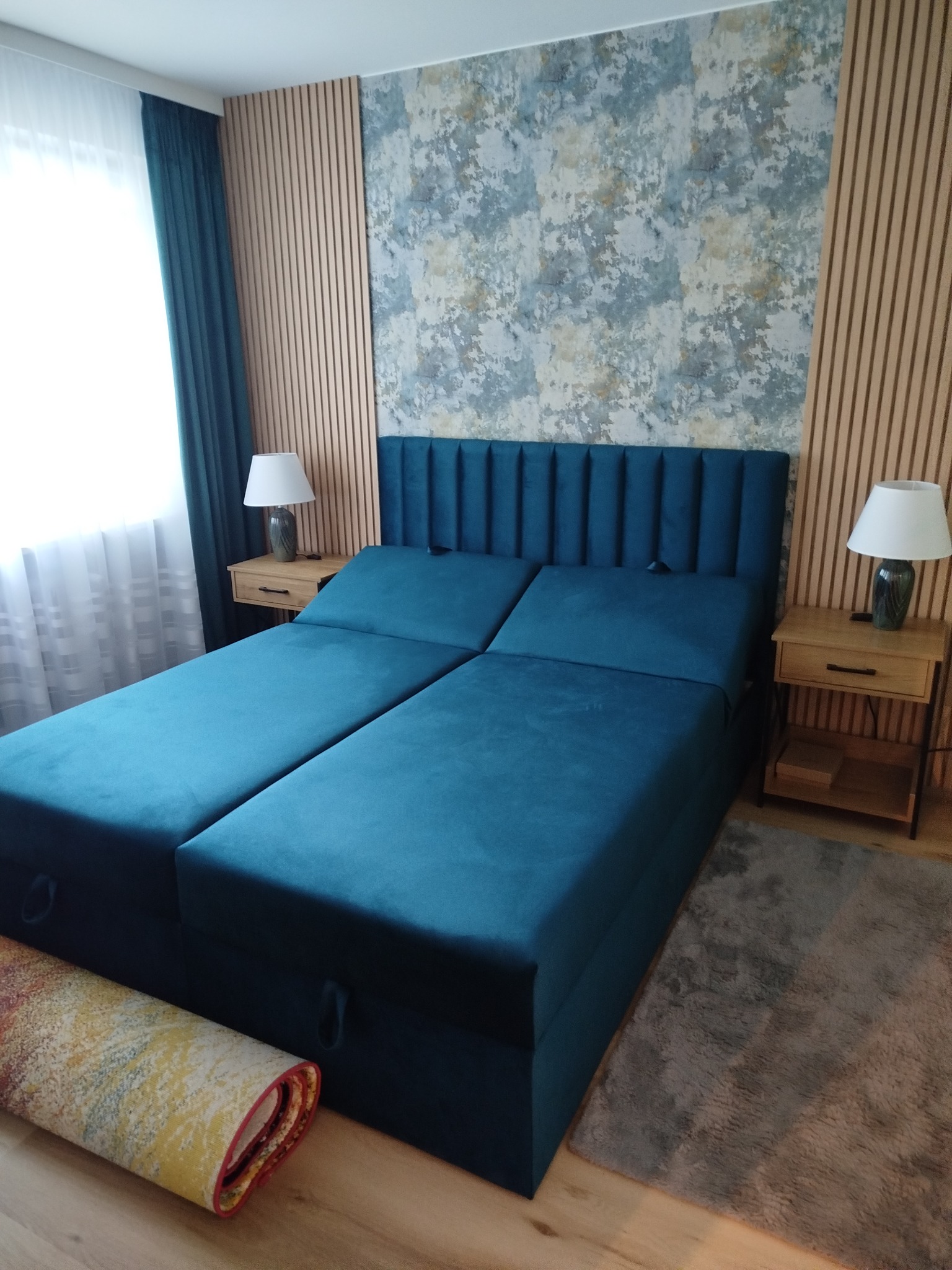 Nowoczesna sypialnia z turkusowym łóżkiem kontynentalnym, drewnianymi lamelami na ścianie i wzorzystą tapetą. Widoczne szafki nocne z lampkami.