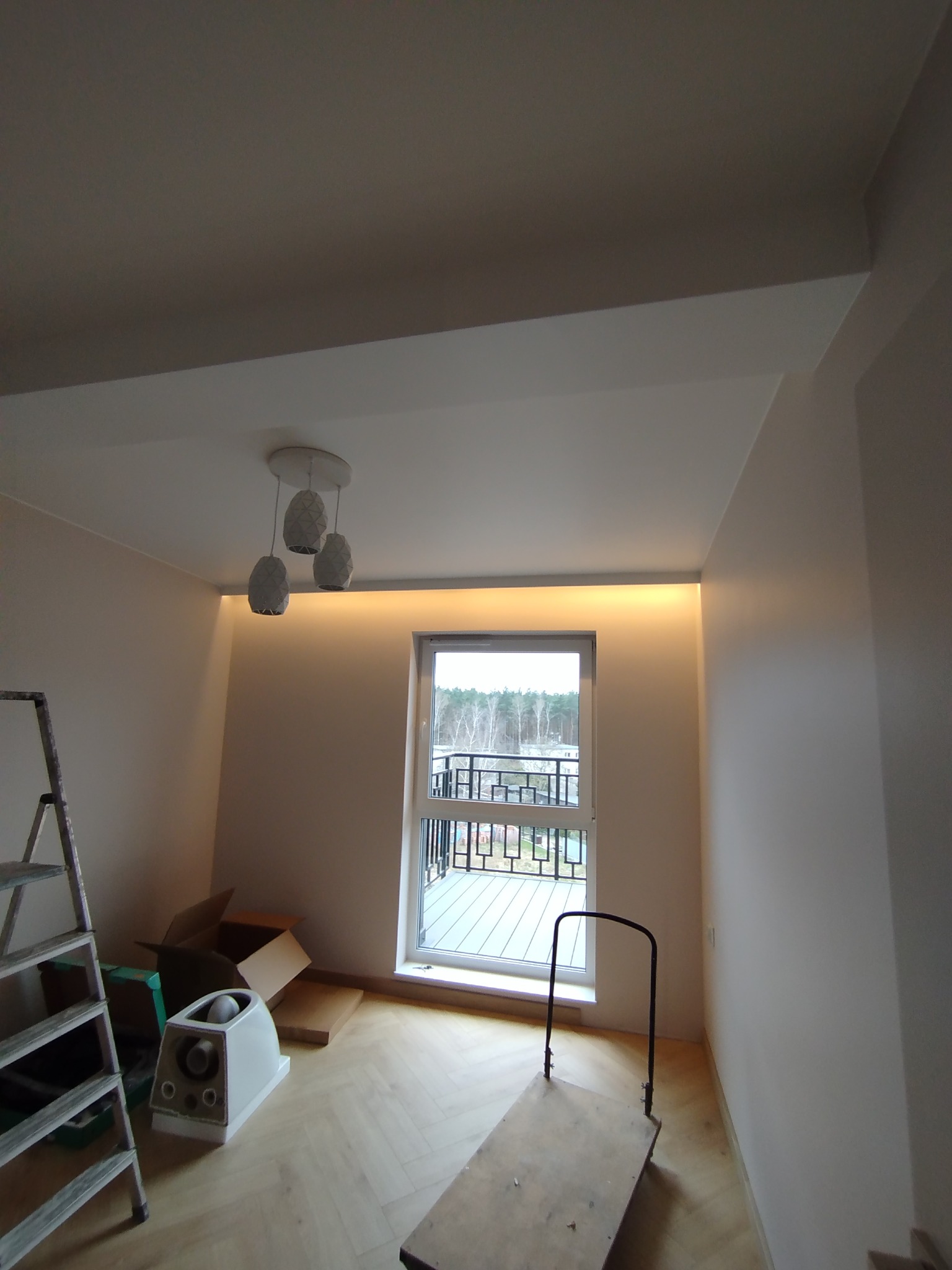 Jasne wnętrze po remoncie z oświetleniem LED podsufitowym, drabina i wózek transportowy. Widoczny parkiet w jodełkę i okno z widokiem na balkon.