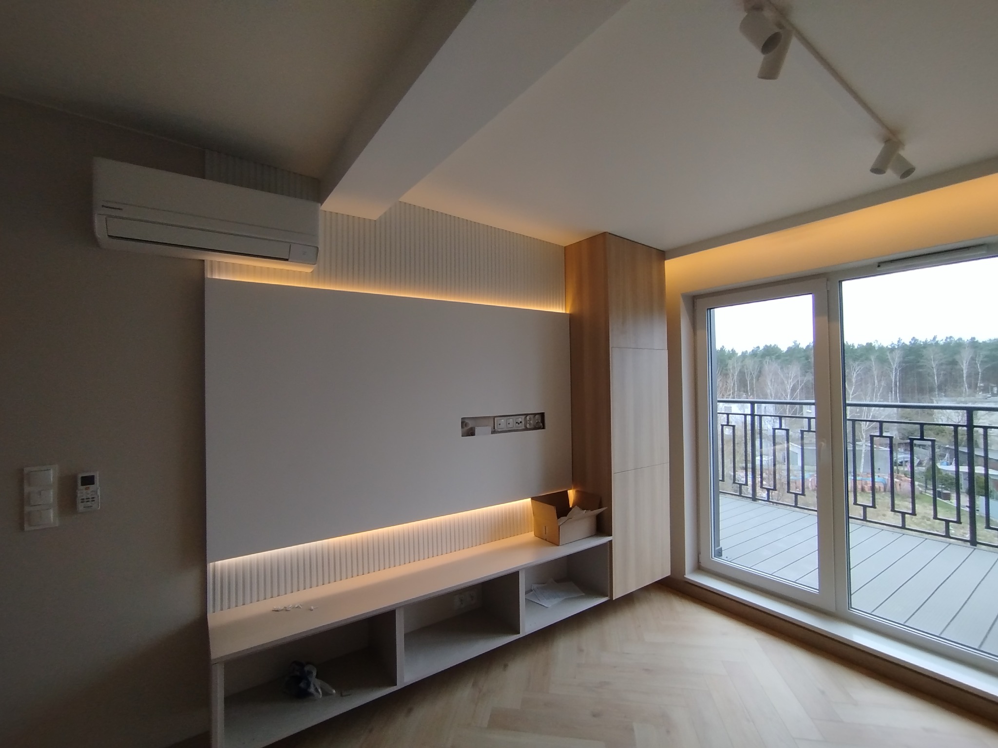 Nowoczesny salon z klimatyzacją, oświetleniem LED i zabudową meblową z drewna. Widok na balkon przez duże okno. Parkiet w jodełkę na podłodze.