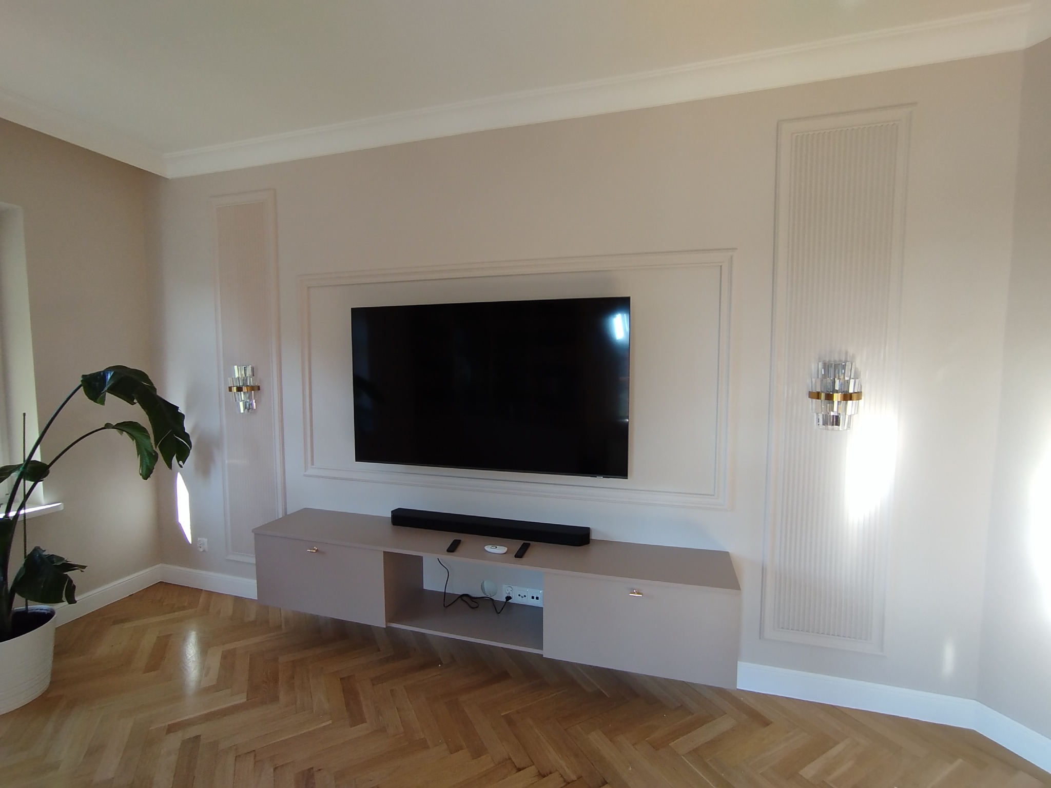 Elegancki salon z telewizorem na ścianie, pod nim szafka RTV. Na ścianach dekoracyjne panele i lampy. Parkiet w jodełkę na podłodze. Minimalistyczny design i stonowana kolorystyka.