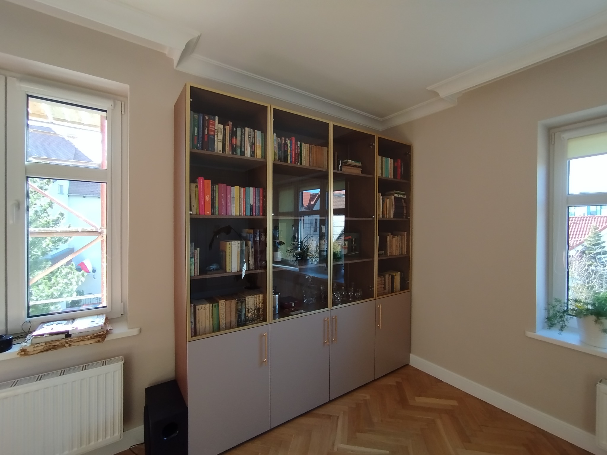 Zabudowa biblioteczna z przeszklonymi witrynami i szarymi szafkami, widok na parkiet w jodełkę, dekoracyjne listwy przysufitowe. Jasne wnętrze z oknami.