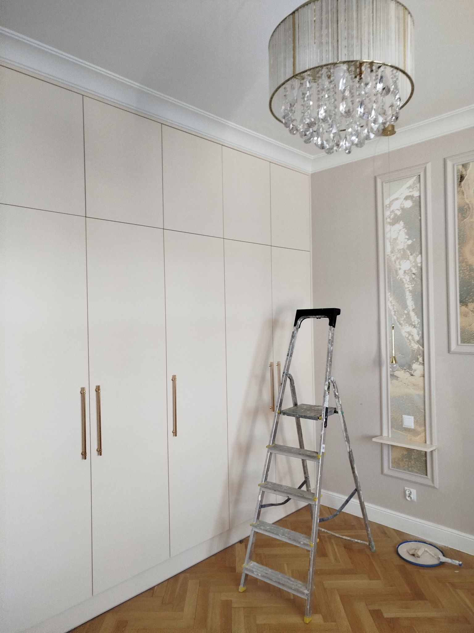 Wnętrze pokoju po wykończeniu: kremowa zabudowa szafy z złotymi uchwytami, drabina malarska, parkiet w jodełkę i ozdobny żyrandol.