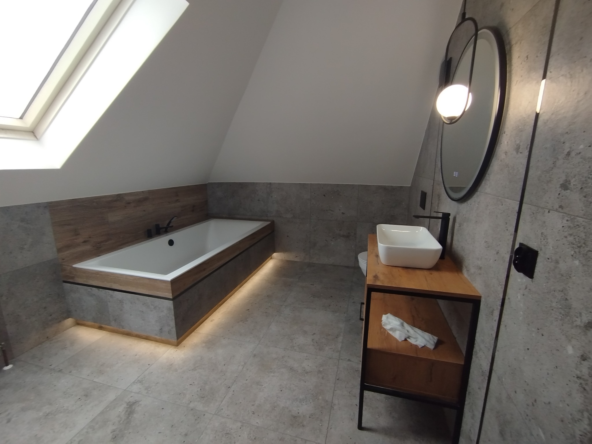 Nowoczesna łazienka na poddaszu z wanną w zabudowie, umywalką na szafce z drewnianymi akcentami i okrągłym lustrem z podświetleniem.