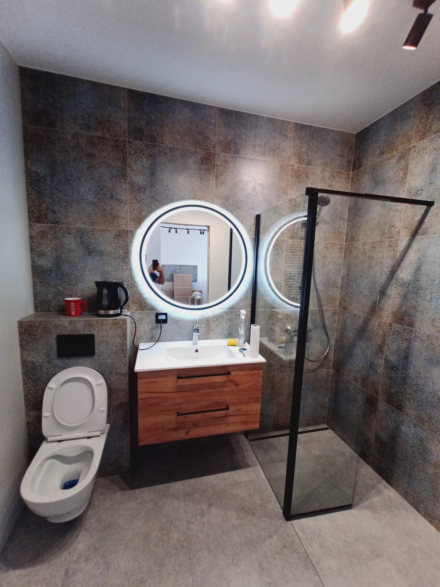 Nowoczesna łazienka z kabiną prysznicową typu walk-in, umywalką z drewnianą szafką i okrągłym lustrem z podświetleniem LED. Na ścianach płytki imitujące kamień.