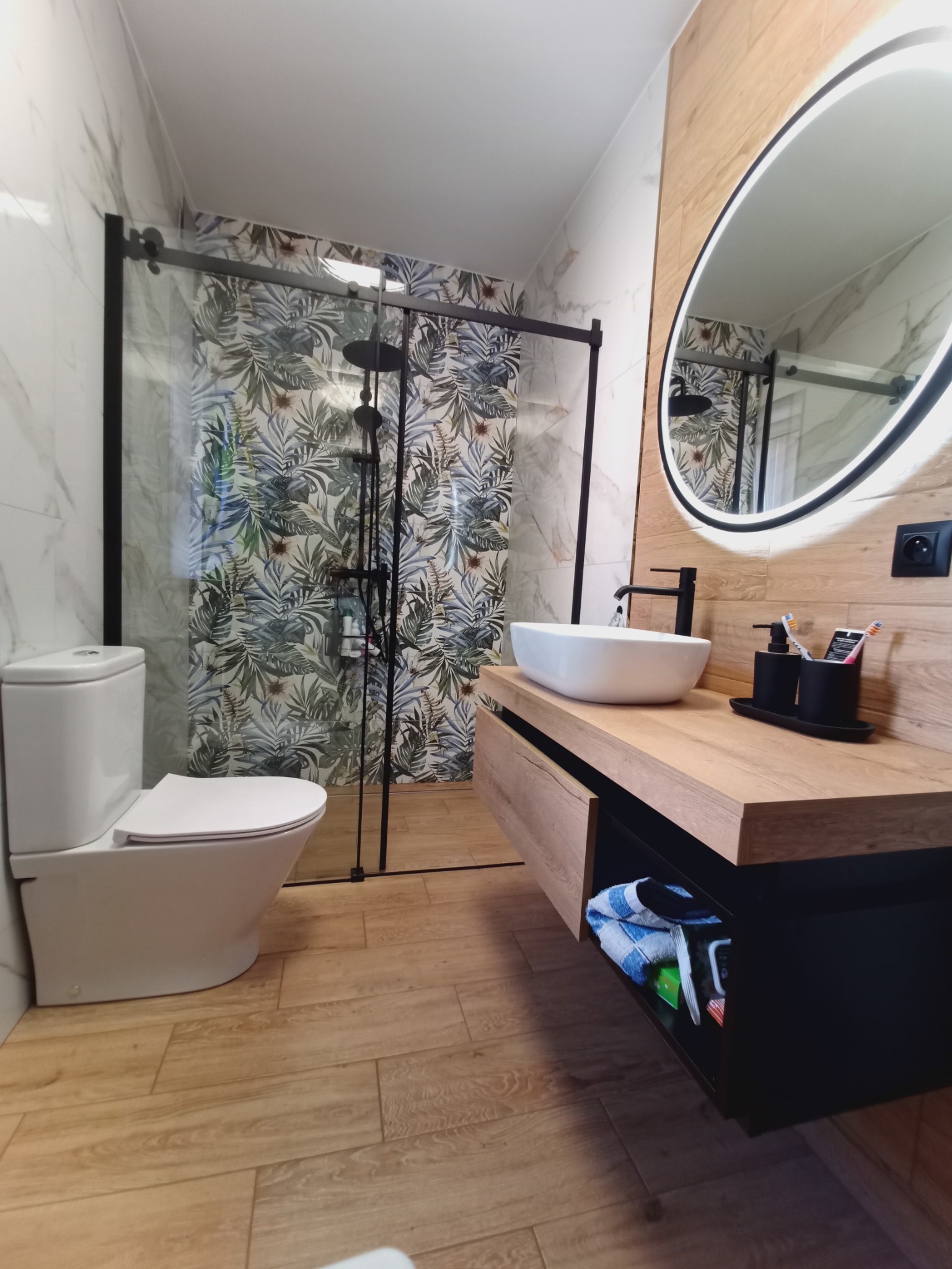 Nowoczesna łazienka w Czarnowie: kabina prysznicowa z motywem roślinnym, umywalka nablatowa i podświetlane lustro owalne. Drewnopodobne płytki i czarne akcenty.
