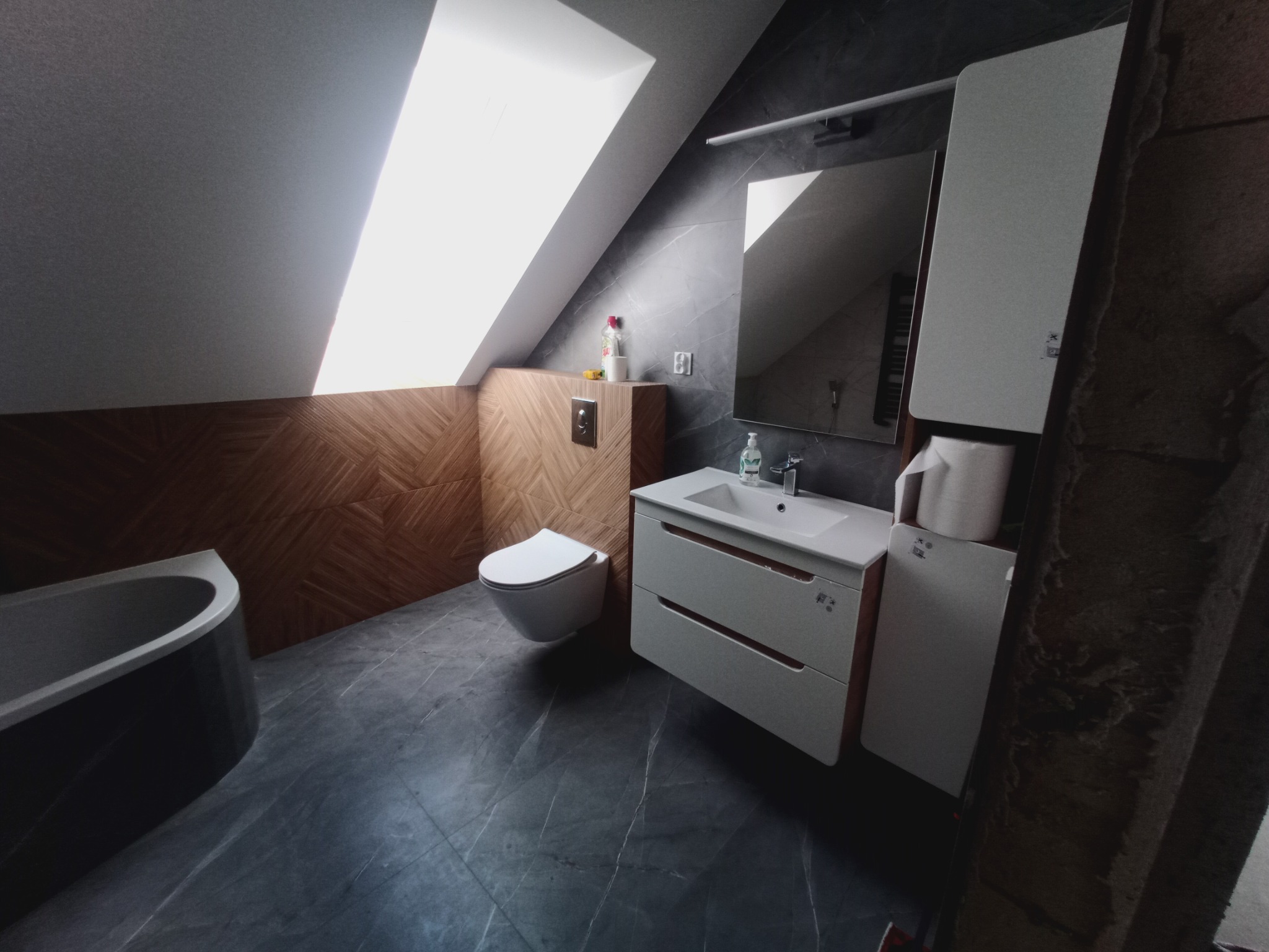 Nowoczesna łazienka na poddaszu z drewnianą boazerią, marmurową podłogą i białymi meblami. Skośny sufit z oknem dachowym. Widok na wannę, toaletę i umywalkę.