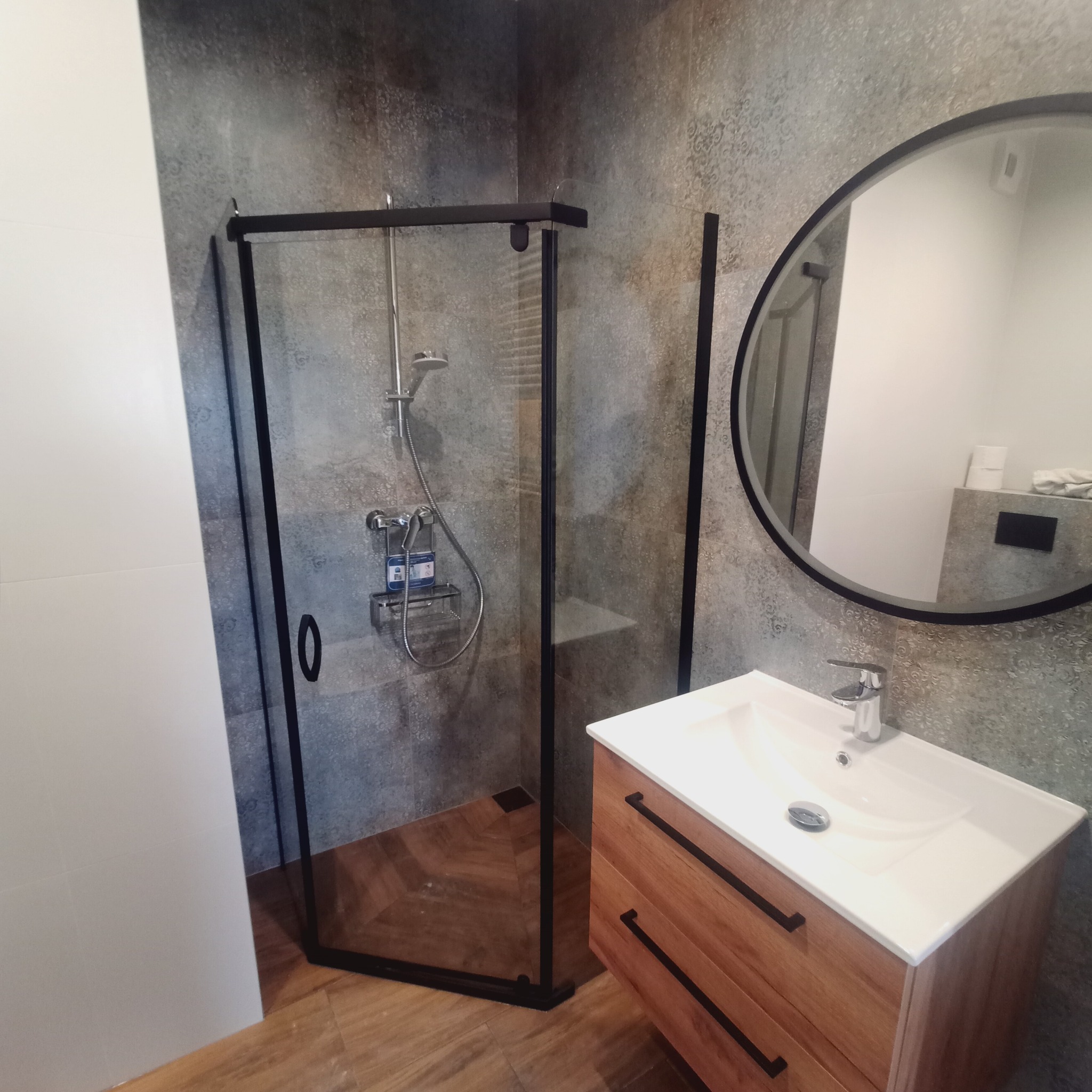 Nowoczesna łazienka z kabiną prysznicową typu walk-in z czarną ramą, umywalką z drewnianą szafką i okrągłym lustrem na tle ściany wykończonej płytkami w odcieniach szarości.