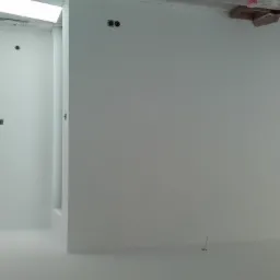 Wnętrze w trakcie prac wykończeniowych: białe ściany i podłoga, widoczne puszki elektryczne, fragment okna z czarną ramą i elementy konstrukcyjne pod sufitem.