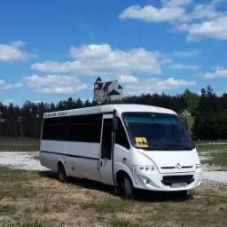 Biały minibus z żółtą naklejką z przodu, zaparkowany na trawiastym terenie z widokiem na zamek w tle pod błękitnym niebem z chmurami.