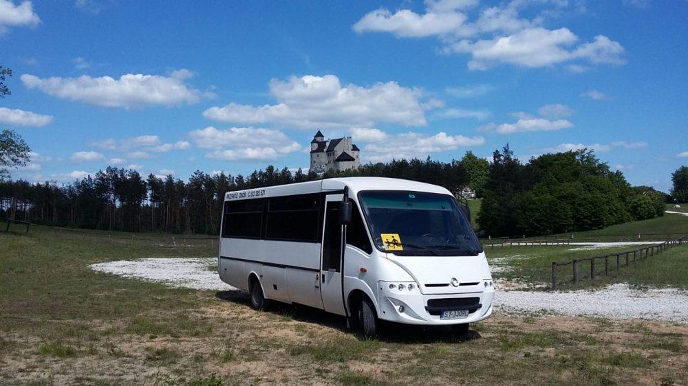 Biały minibus z żółtą naklejką z przodu, zaparkowany na trawiastym terenie z widokiem na zamek w tle pod błękitnym niebem z chmurami.