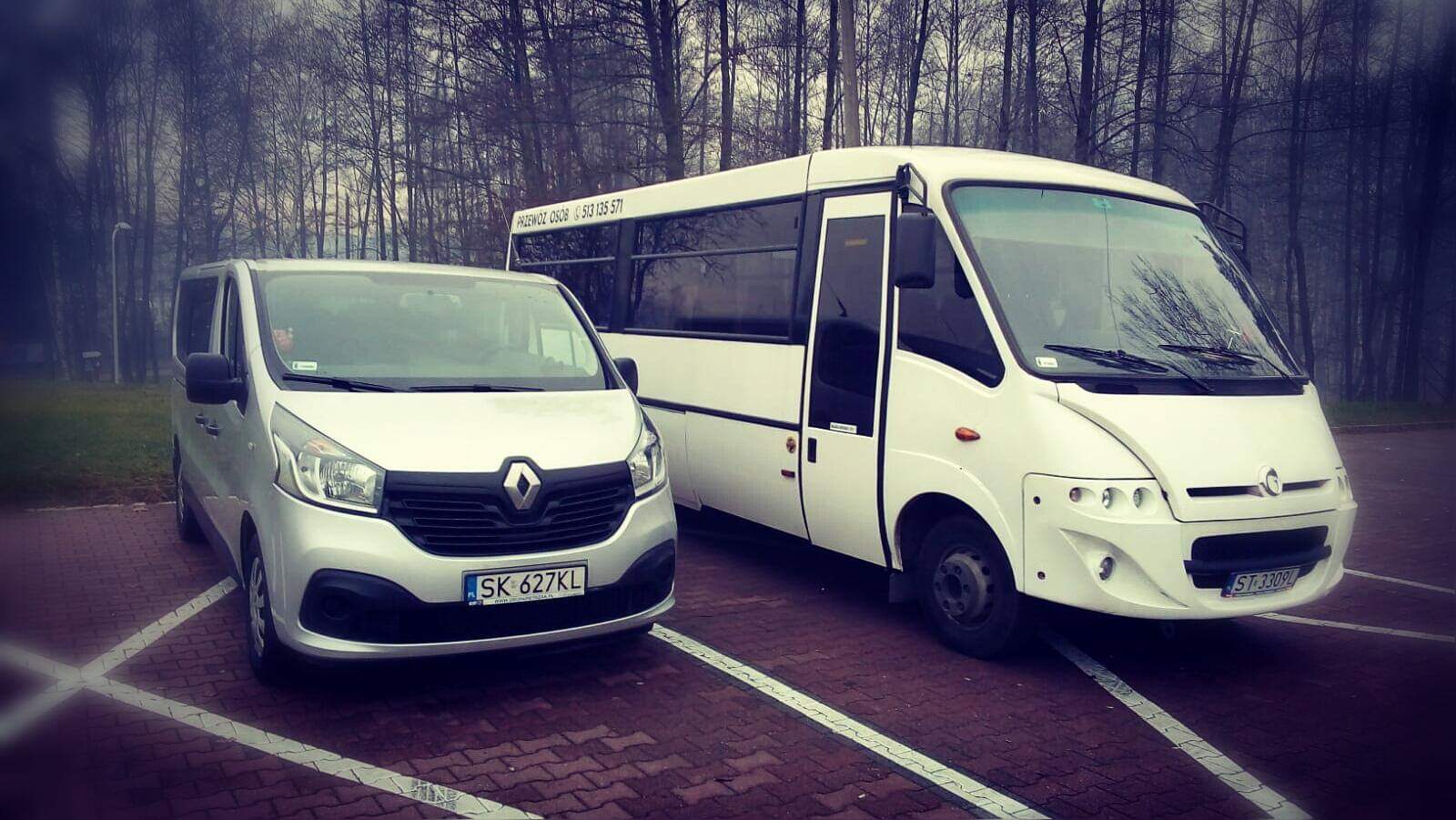 Dwa busy: srebrny Renault Trafic z przodu i biały autobus z numerem telefonu na dachu, stoją zaparkowane na kostce brukowej w wyznaczonym miejscu, w tle drzewa bez liści.