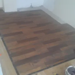 Proces układania ciemnobrązowych paneli podłogowych w pomieszczeniu, widoczne niedokończone fragmenty, narzędzia i wiadro z klejem w rogu.