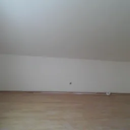 Poddasze po remoncie: świeżo pomalowane ściany w odcieniach bieli, drewniana podłoga, widoczne gniazdko elektryczne na ścianie, oraz fragment okna z zabezpieczeniami.