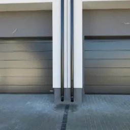 Dwie szare bramy segmentowe przemysłowe z poziomymi przetłoczeniami, oddzielone pionową kolumną z dwiema rurami spustowymi, widok z zewnątrz na szarym budynku z żółtą skrzynką gazową po lewej stronie.