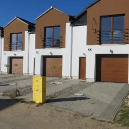 Szereg nowych domów jednorodzinnych z drewnopodobnymi bramami garażowymi, oknami i balkonami, żółte skrzynki gazowe przed budynkami.