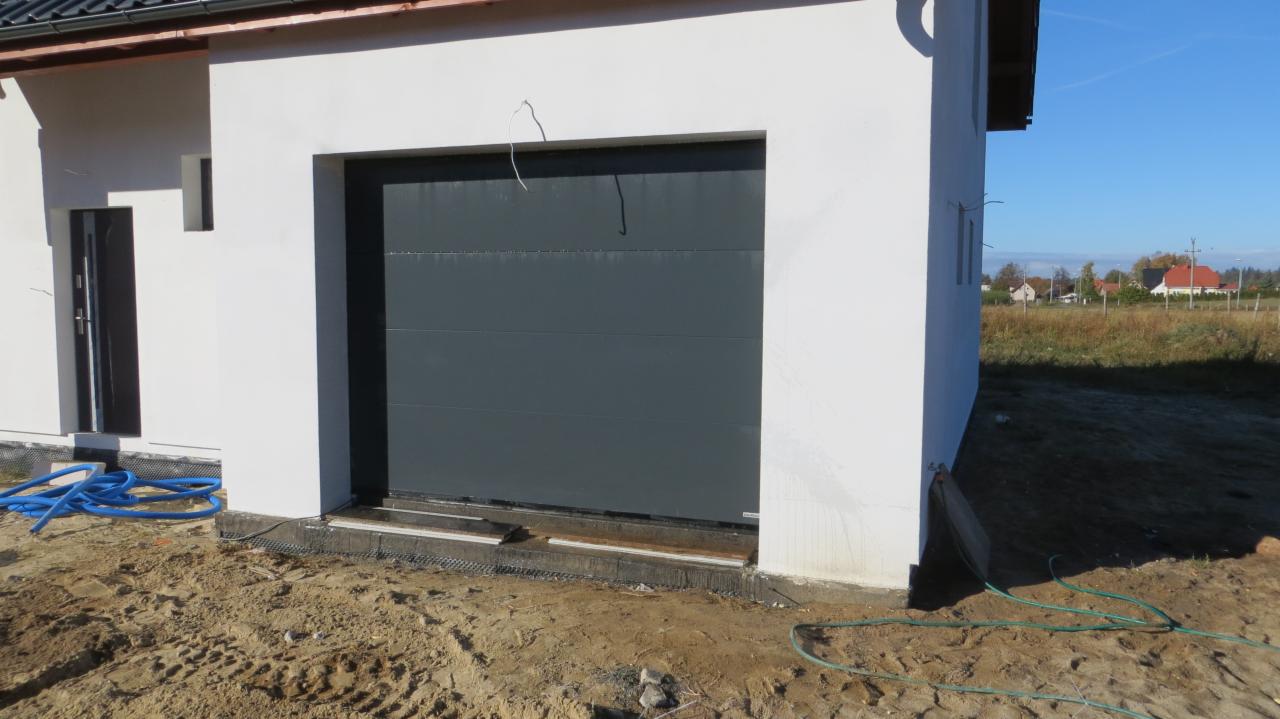 Szara brama garażowa segmentowa w nowym budynku z białą elewacją, widoczne niedokończone prace ziemne i niebieski wąż doprowadzający wodę.