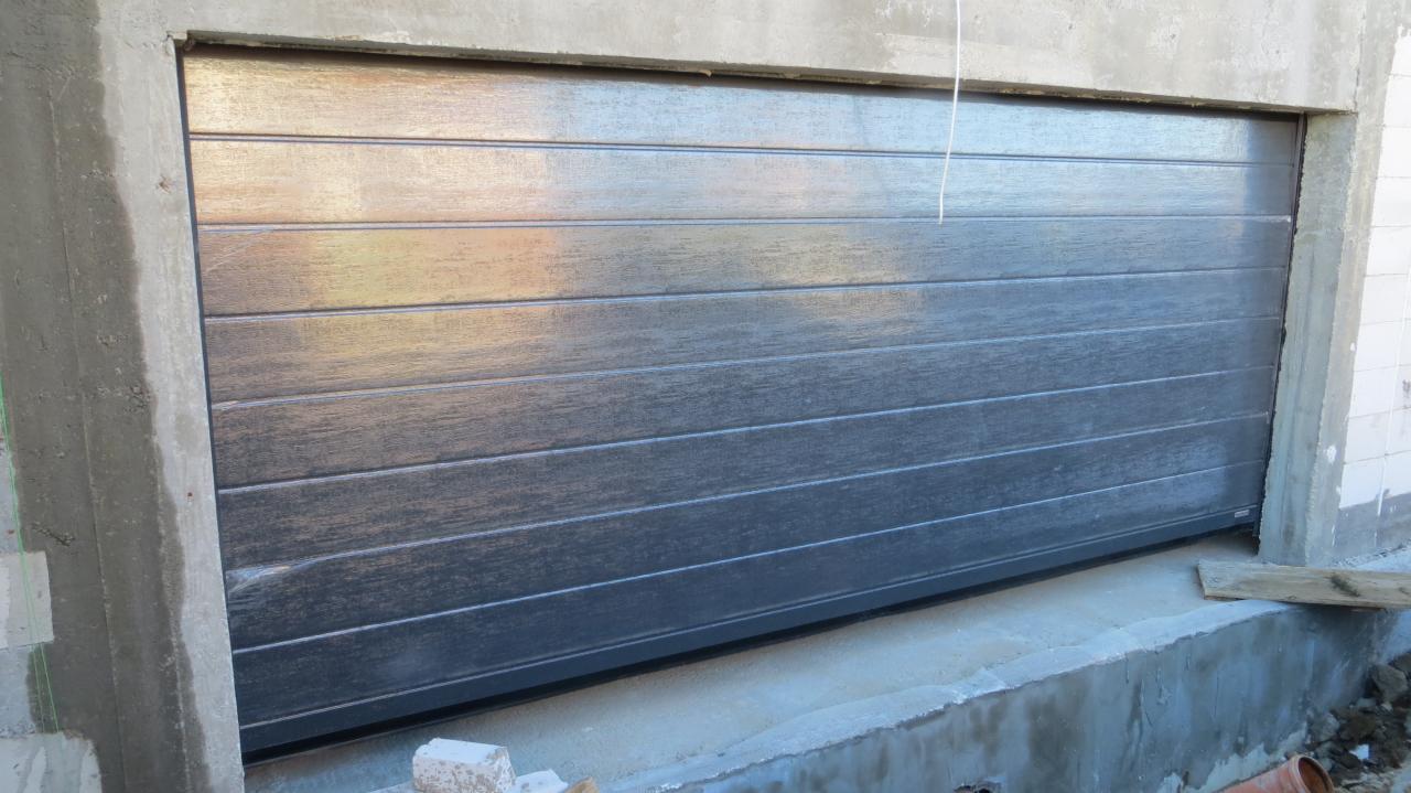 Zamknięta, pozioma brama garażowa w kolorze szaro-brązowym, z widocznymi poziomymi panelami, osadzona w surowej, betonowej ramie podczas prac budowlanych.