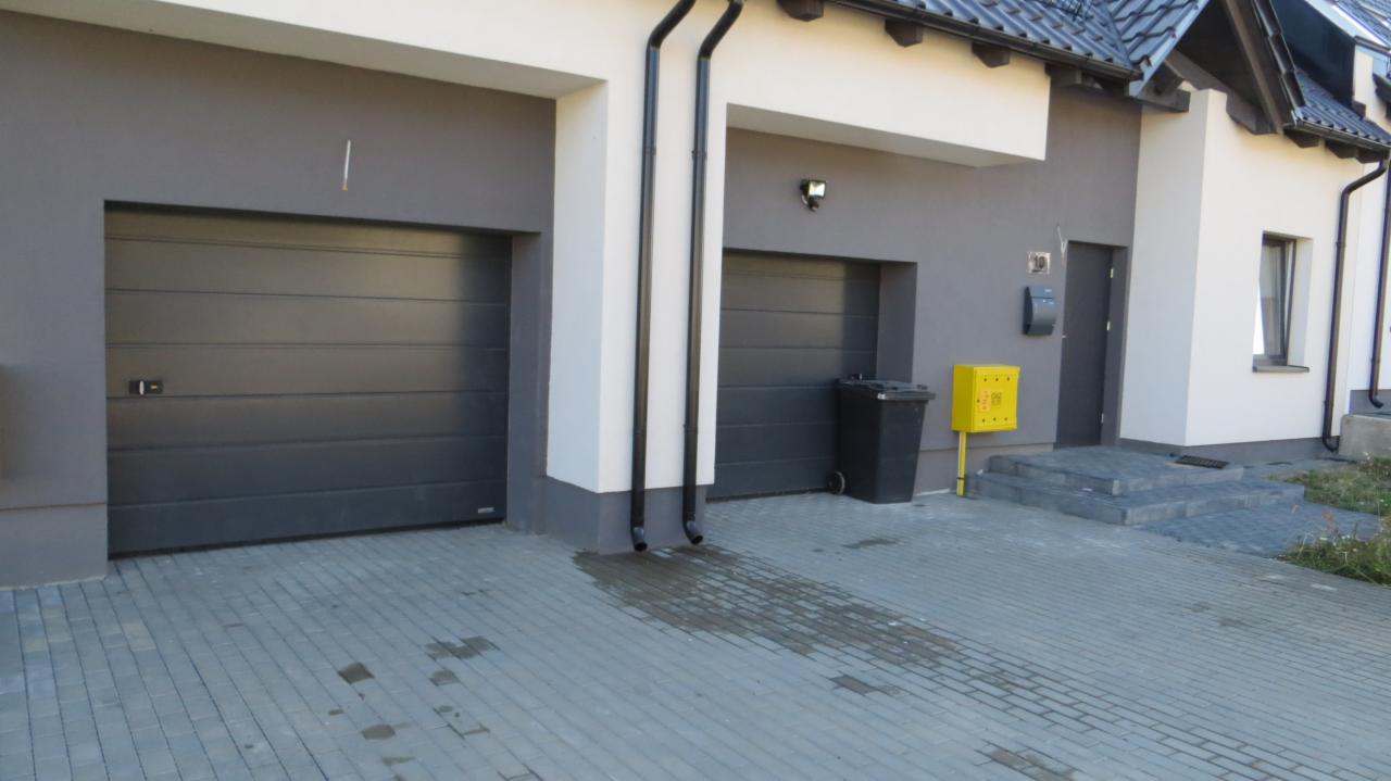 Dwa garaże z szarymi bramami segmentowymi, czarne rynny spływowe, kostka brukowa na podjeździe, skrzynka gazowa w kolorze żółtym.