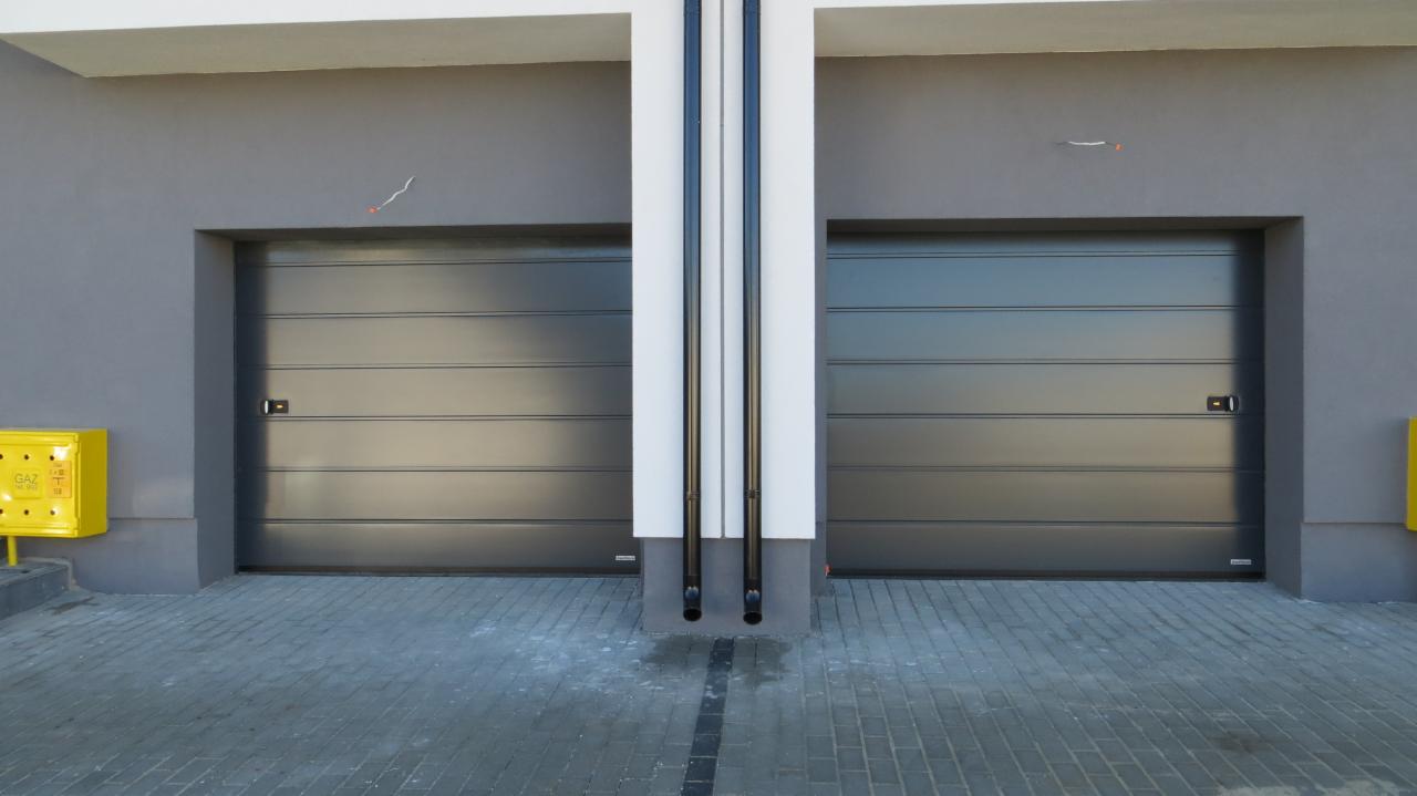 Dwie szare bramy segmentowe przemysłowe z poziomymi przetłoczeniami, oddzielone pionową kolumną z dwiema rurami spustowymi, widok z zewnątrz na szarym budynku z żółtą skrzynką gazową po lewej stronie.