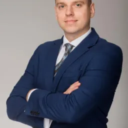 Portret mężczyzny w niebieskim garniturze z rękami skrzyżowanymi na tle szarego tła, z widocznym wzorem na krawacie.