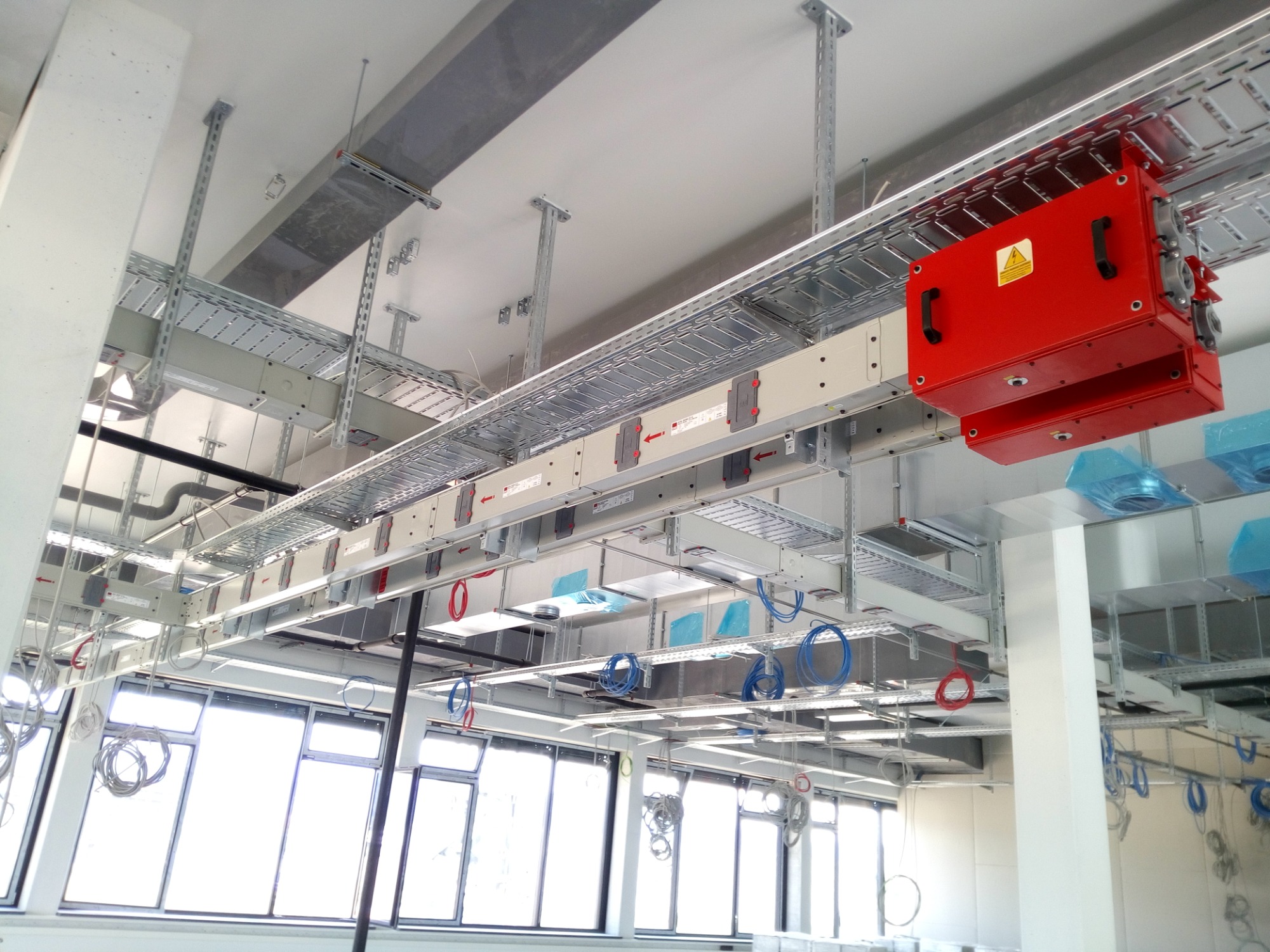 Widok sufitu w hali produkcyjnej z zamontowanymi korytkami kablowymi, przewodami i czerwoną skrzynką elektryczną z symbolem ostrzegawczym.
