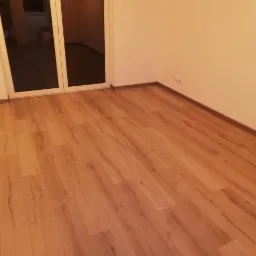 Puste pomieszczenie z nową, drewnopodobną podłogą i świeżo pomalowanymi ścianami w odcieniu ciepłej bieli, widoczne gniazdka elektryczne przy podłodze, duże przeszklone drzwi balkonowe.