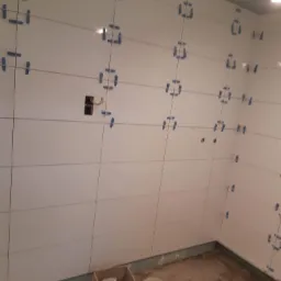 Świeżo ułożone, białe, poziome płytki w łazience, zabezpieczone niebieską taśmą dystansową, widoczne puszki elektryczne, podłoga w trakcie przygotowania.