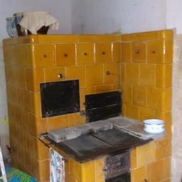 Ciepło i Styl Robert Karczmarski - Żółty piec kaflowy z czarnymi drzwiczkami w rogu pomieszczenia, z widocznymi akcesoriami kuchennymi i fragmentem drabiny.