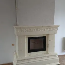 Wykończony, kremowy kominek kaflowy z czarną wkładką, zdobiony ornamentami, w surowym wnętrzu z widocznym gniazdkiem elektrycznym i fragmentem grzejnika.