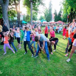 Grupa dzieci uczestniczy w zabawie z niskim limbo na trawie podczas imprezy plenerowej, w tle widoczni dorośli, trampolina i budynki.