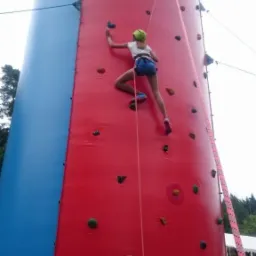 Dziewczynka w kasku wspina się po dmuchanej, czerwono-niebieskiej ściance wspinaczkowej z różnokolorowymi uchwytami, zabezpieczona liną.