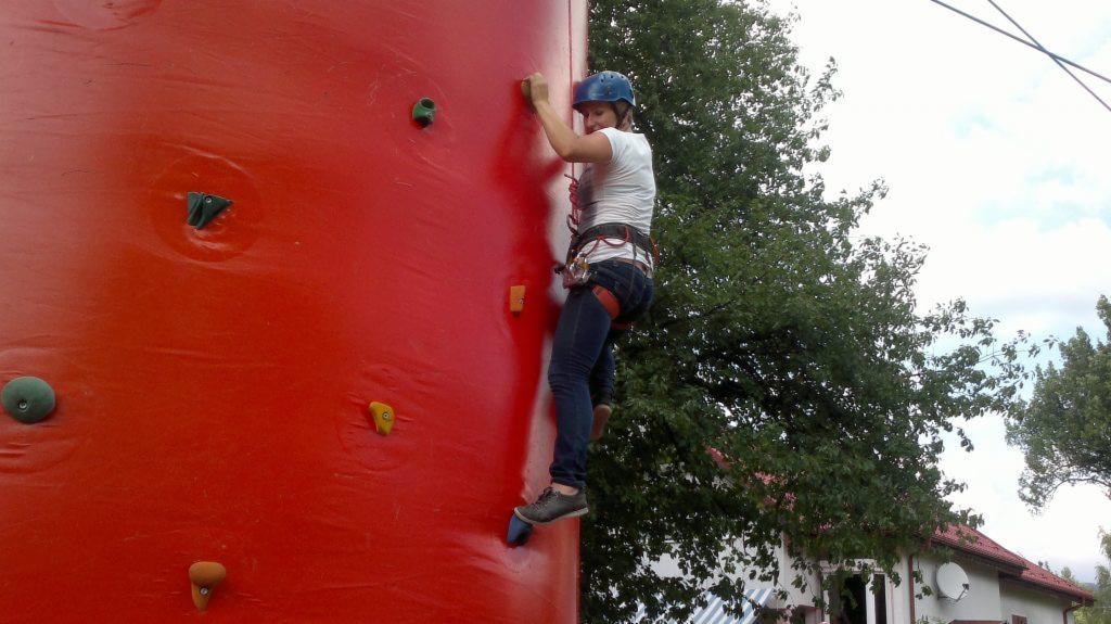 Kobieta w kasku i uprzęży wspinaczkowej wspina się po czerwonej ściance wspinaczkowej z różnokolorowymi chwytami na zewnątrz.
