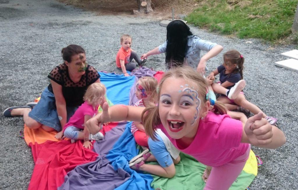 Grupa dzieci i dorosłych z pomalowanymi twarzami bawi się na kolorowej chuście na żwirowym podłożu, dziewczynka w różowej koszulce uśmiecha się szeroko, pokazując kciuk w górę.