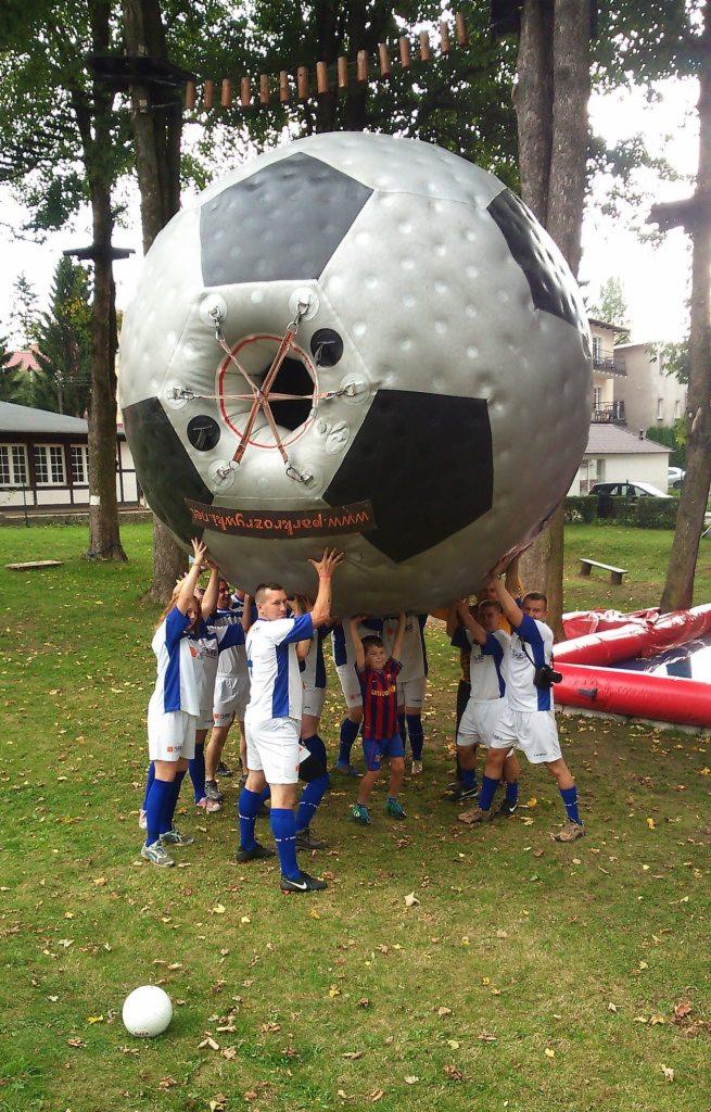 Grupa osób w Kudowie-Zdroju podtrzymuje dużą, dmuchaną piłkę nożną z widocznym napisem www.parkrozrywki.pl. W tle drzewa i budynki.