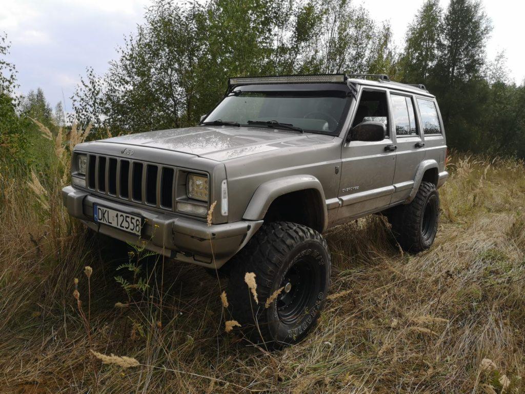 Szary Jeep Cherokee z oponami terenowymi i dodatkowym oświetleniem LED na dachu, zaparkowany na łące z wysoką trawą, tablica rejestracyjna DKL 12558.