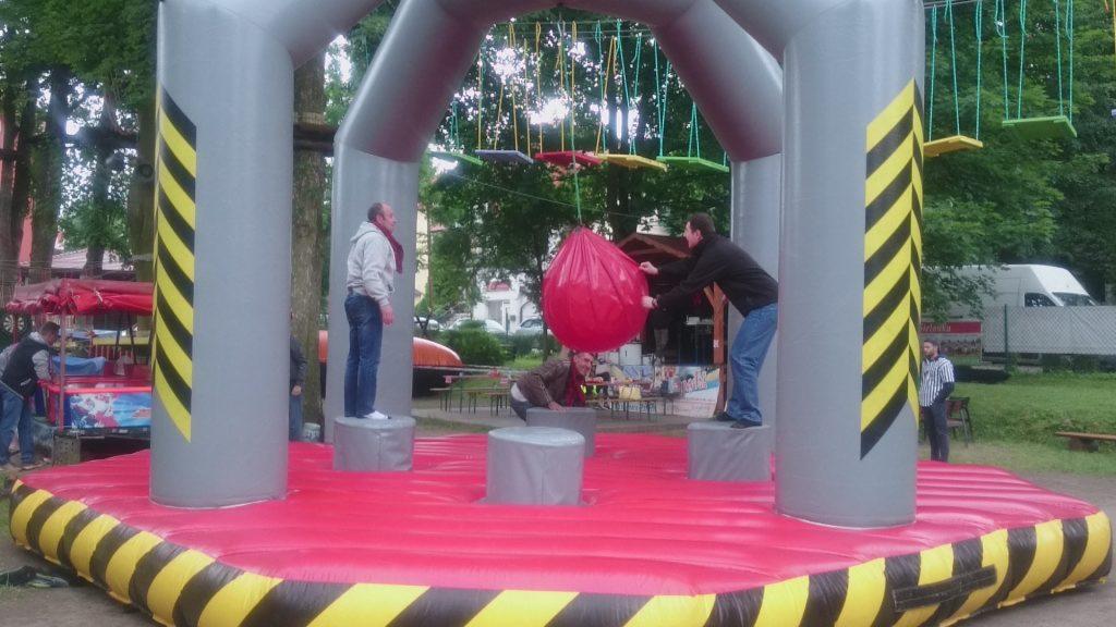 Uczestnicy imprezy integracyjnej na dmuchanym torze przeszkód z wiszącą czerwoną kulą do zbijania, widoczne osoby w różnym wieku i strojach, tło z drzewami i budynkami.