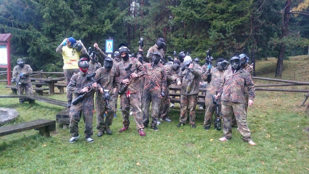Grupa osób w strojach kamuflażowych i maskach ochronnych, trzymających markery do paintballa, pozuje na tle leśnym z ławkami i stołami piknikowymi.