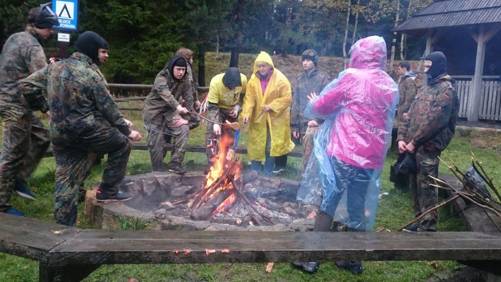 Grupa osób w różnorodnych ubraniach, w tym w strojach moro i płaszczach przeciwdeszczowych, zgromadzona wokół ogniska w kamiennym kręgu, na którym pieką kiełbaski, na tle leśnego krajobrazu.
