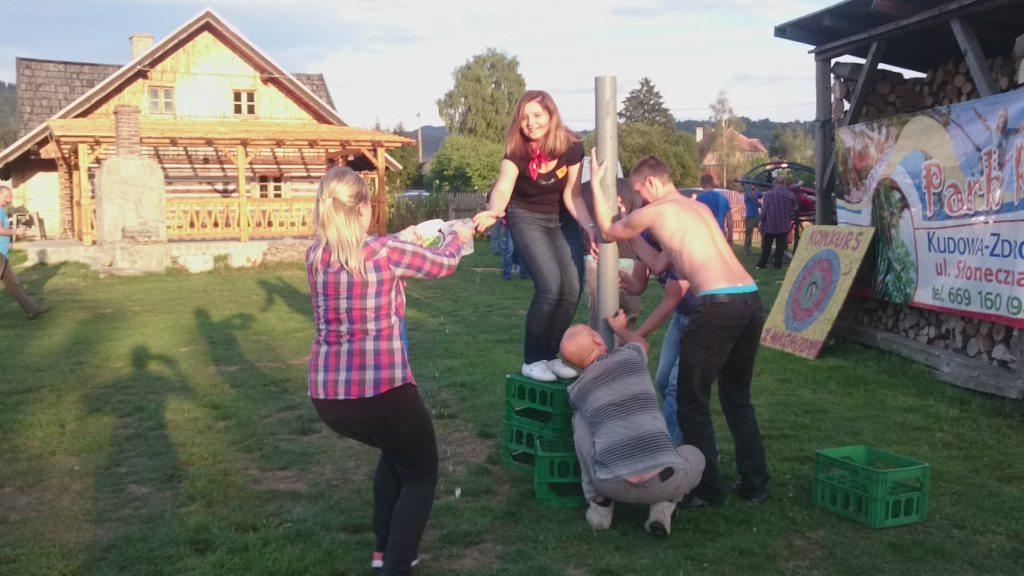 Grupa osób uczestniczy w zabawie plenerowej z użyciem rury i skrzynek, w tle drewniany budynek i baner z informacjami o konkursie.