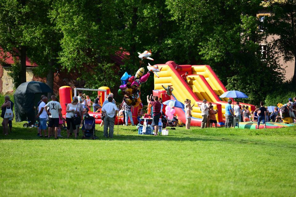 Dmuchana zjeżdżalnia w kolorach żółto-czerwonych na zielonej trawie, otoczona bawiącymi się dziećmi i rodzicami, z balonami w kształcie samolotów i kolorowymi balonami na pierwszym planie.
