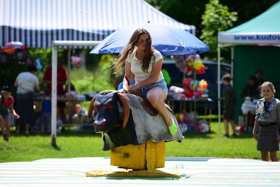 Młoda kobieta w dżinsowych szortach i jasnozielonych skarpetkach siedzi na mechanicznym byku na imprezie plenerowej, w tle widoczne kolorowe balony i namioty.