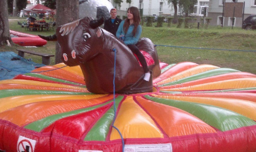 Dziewczyna w niebieskiej bluzie siedzi na dmuchanym, brązowym byku na kolorowej dmuchanej poduszce. W tle widoczni mężczyzna i budynek.