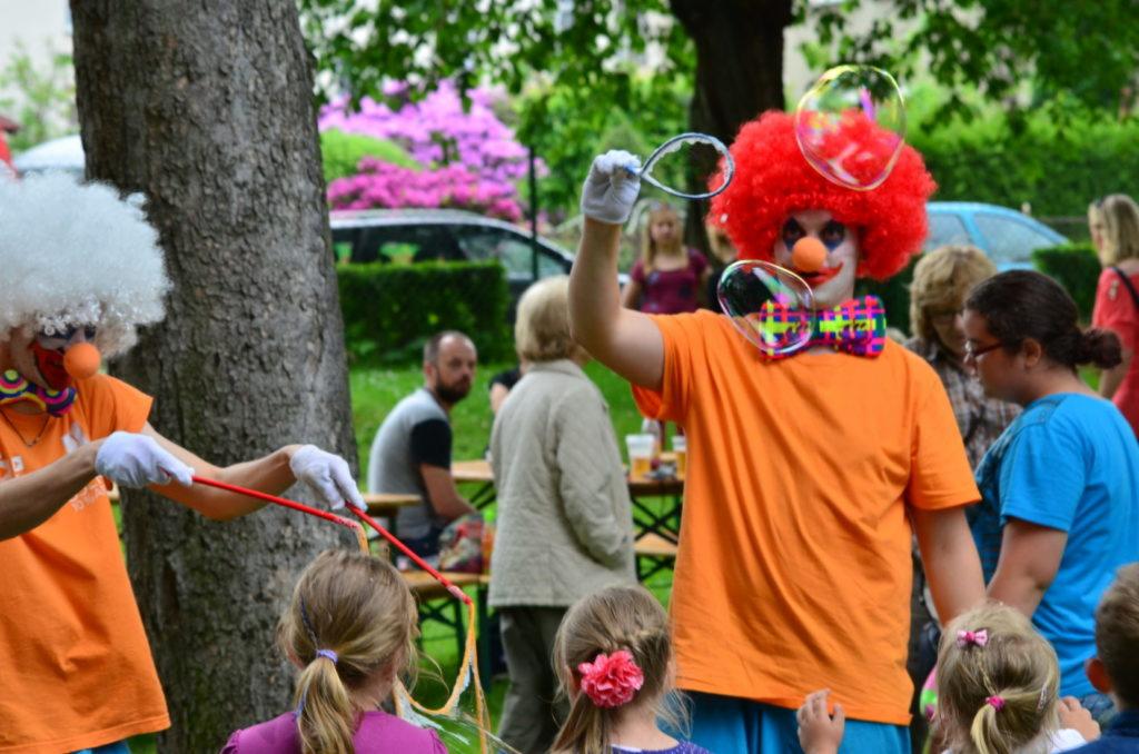 Dwóch klaunów w pomarańczowych koszulkach i białych perukach puszcza bańki mydlane dzieciom na tle drzew i bawiących się ludzi.