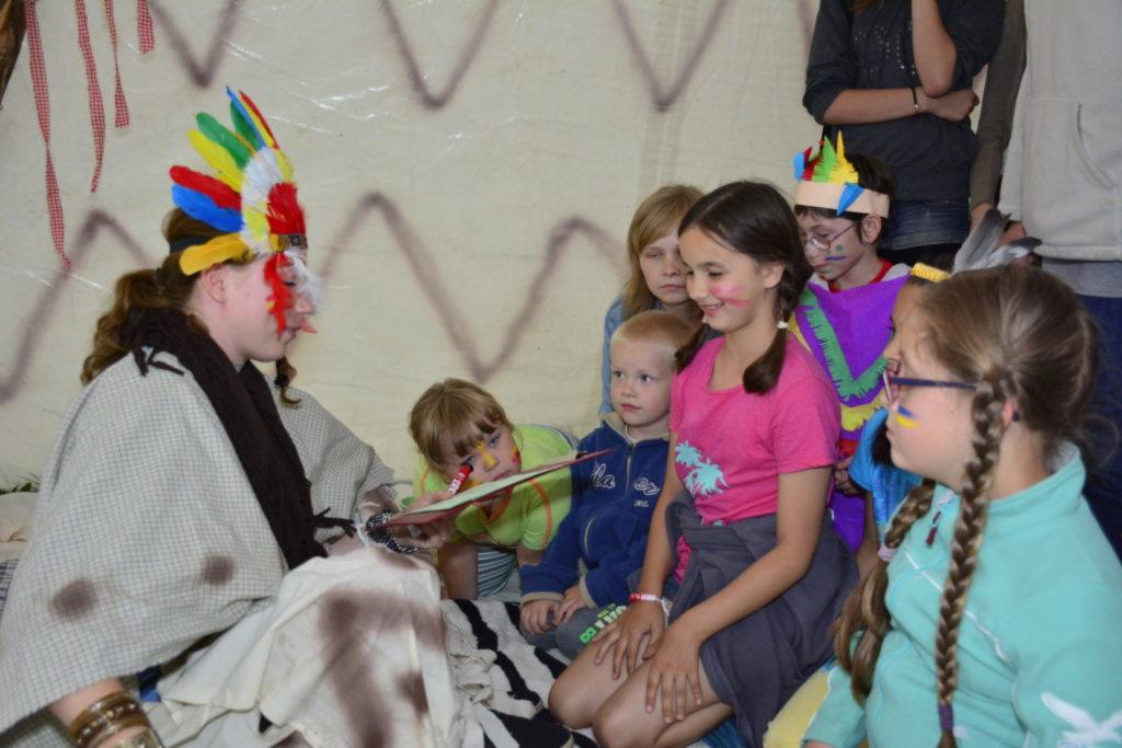 Osoba przebrana za Indianina z pióropuszem na głowie, siedzi na kocu i prowadzi zajęcia dla grupy dzieci w różnym wieku, niektóre z pomalowanymi twarzami, w tle imitacja tipi.