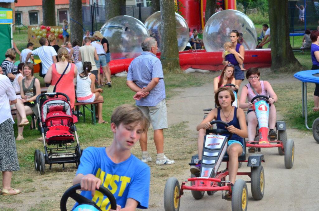 Uczestnicy zabawy na gokartach pedałowych podczas imprezy plenerowej, w tle dmuchane kule i tłum ludzi.