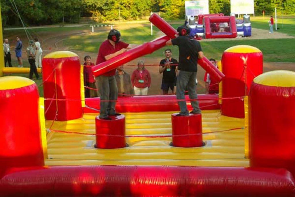Dwaj mężczyźni w kaskach walczą na dmuchanym ringu, używając dmuchanych pałek, publiczność obserwuje w tle.