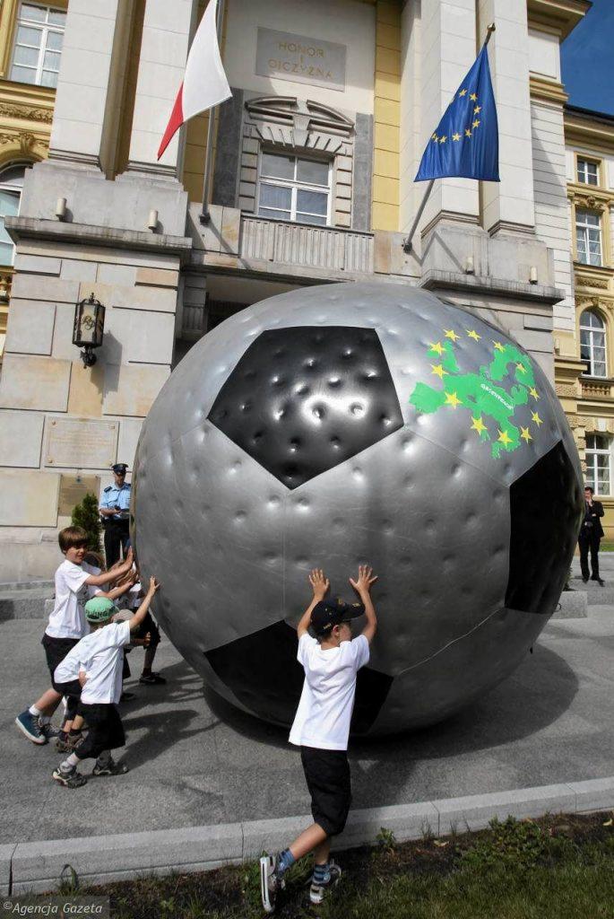 Grupa dzieci próbuje przesunąć ogromną, srebrną piłkę z mapą Europy i gwiazdami, na tle budynku z flagami Polski i Unii Europejskiej.