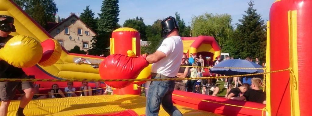 Dmuchany ring bokserski z dwoma uczestnikami w dużych, miękkich rękawicach i ochronnych kaskach, zabawa na świeżym powietrzu podczas imprezy plenerowej.